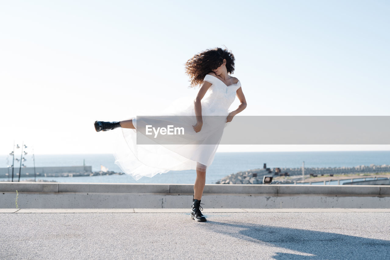 Rebellious unrecognizable female in elegant white dress and brutal boots walking on rooftop in sunlight