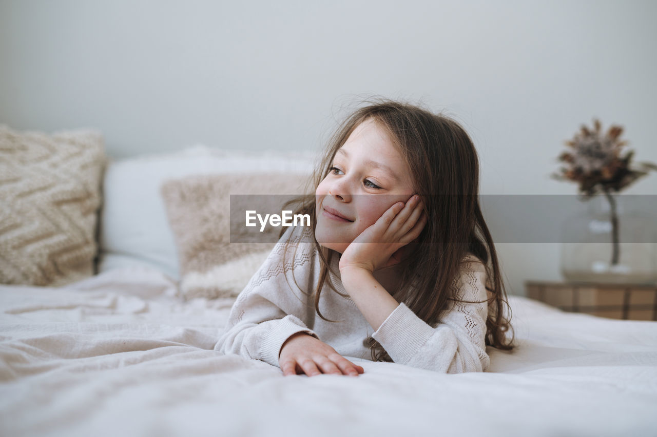 Cuty funny thinking little girl with long hair in home clothes lies on bed at home