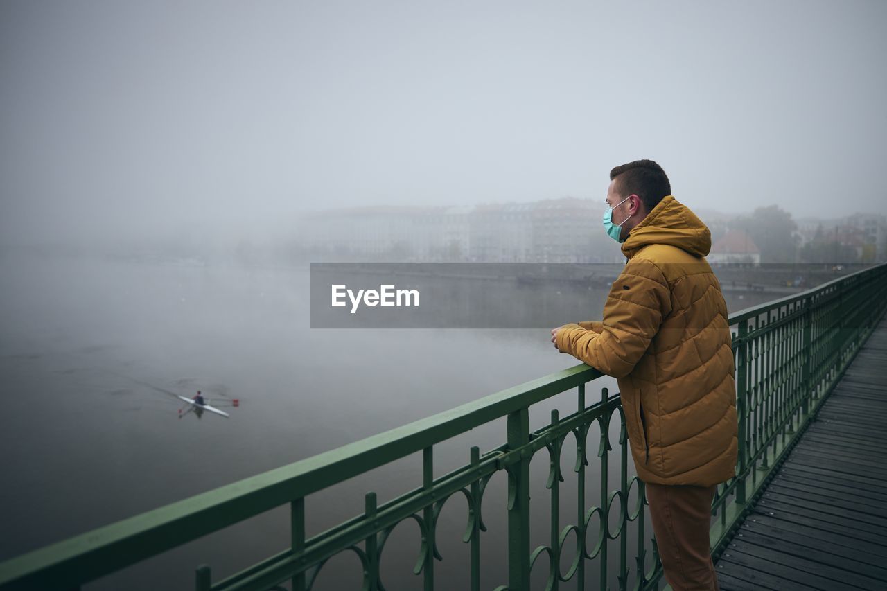 Man wearing mask standing by railing on bridge against sky during winter