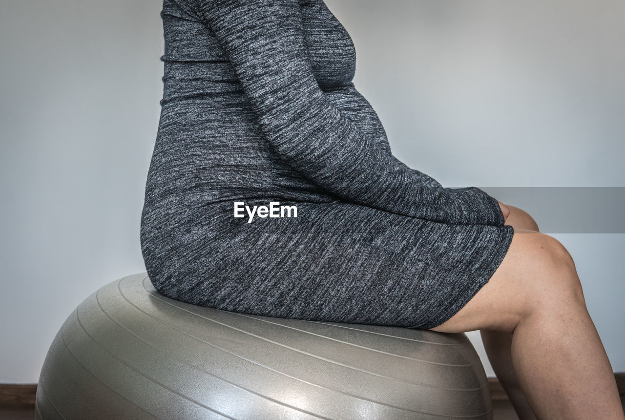 Midsection of pregnant woman sitting on fitness ball against wall