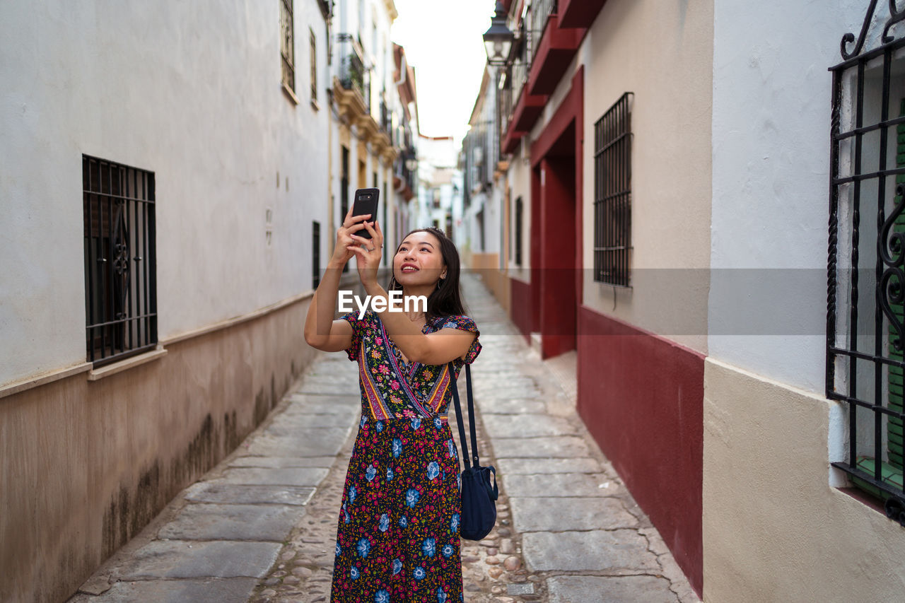 Cheerful asian female tourist taking picture of aged buildings while standing on paved narrow street of cordoba town in spain