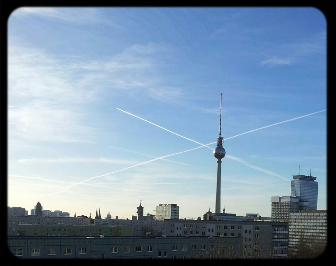 Low angle view of fernsehturm amidst buildings against vapor trails in sky