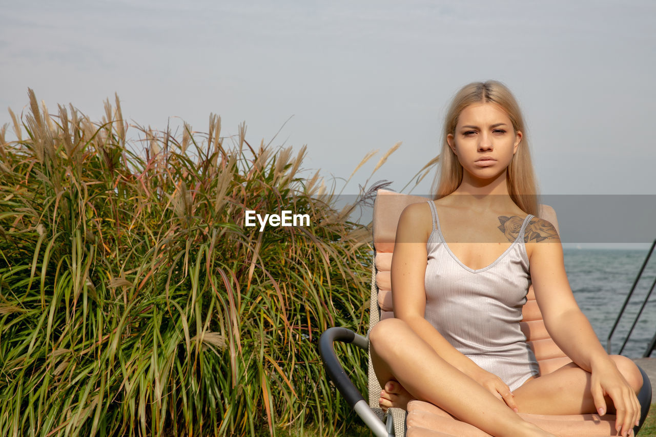 Portrait of beautiful young woman sitting on chair against sea and sky