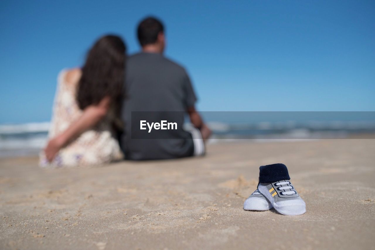 REAR VIEW OF FRIENDS SITTING ON BEACH AGAINST CLEAR BLUE SKY