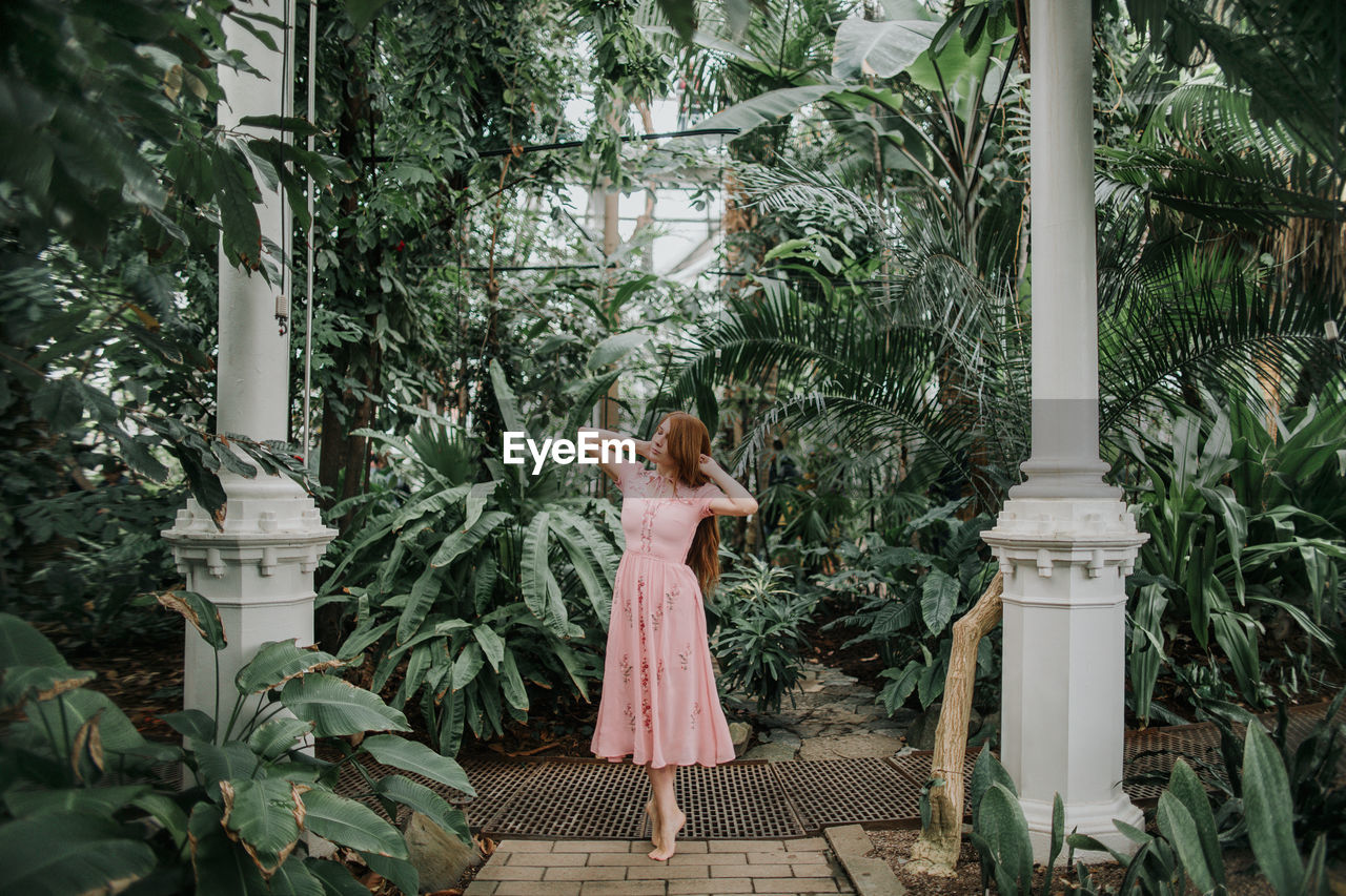 Full body of redhead female standing on tiptoes with hands on head among tropical palm and plants in glasshouse