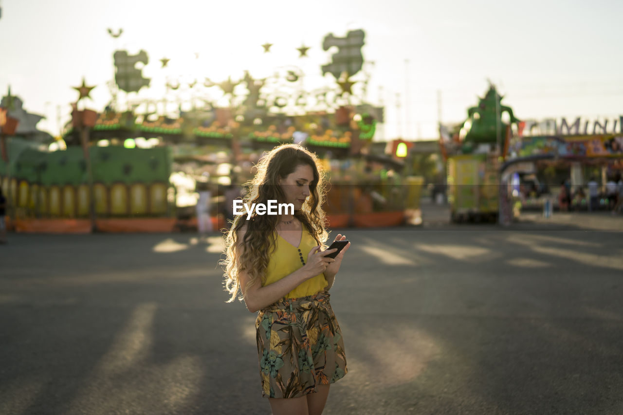 Female with wavy hair standing at fairground and browsing mobile phone at sundown in summer