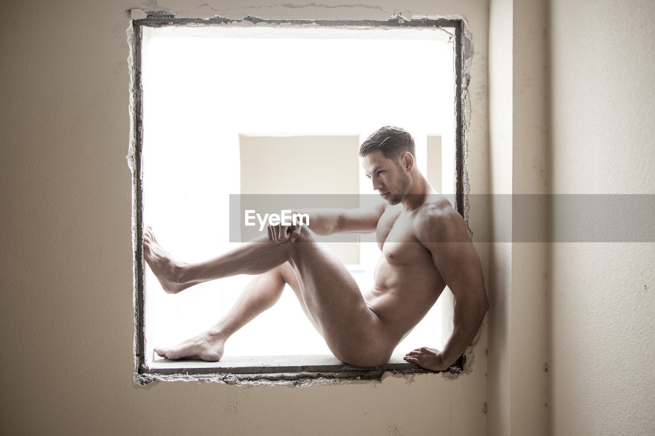 Full length of naked man sitting by window