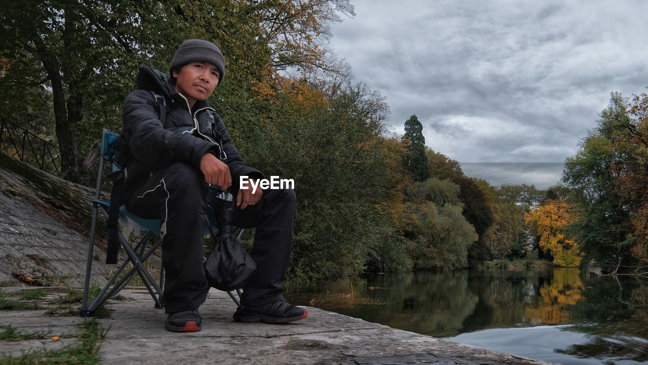 Full length of man sitting by lake against sky during winter