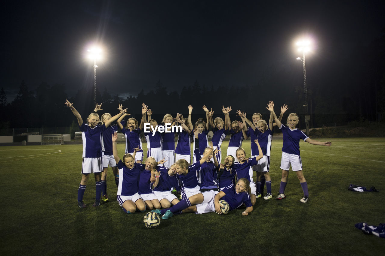 Portrait of cheerful girl's soccer team on illuminated field against sky