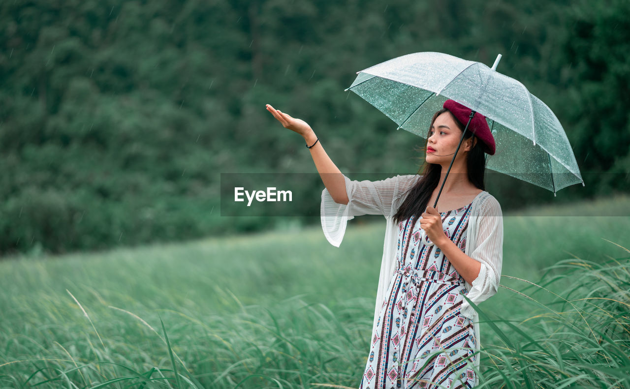 WOMAN HOLDING UMBRELLA WHILE STANDING ON FIELD DURING RAINY DAY