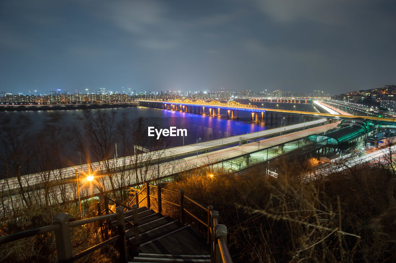 ILLUMINATED BRIDGE OVER RIVER BY CITY AGAINST SKY AT NIGHT