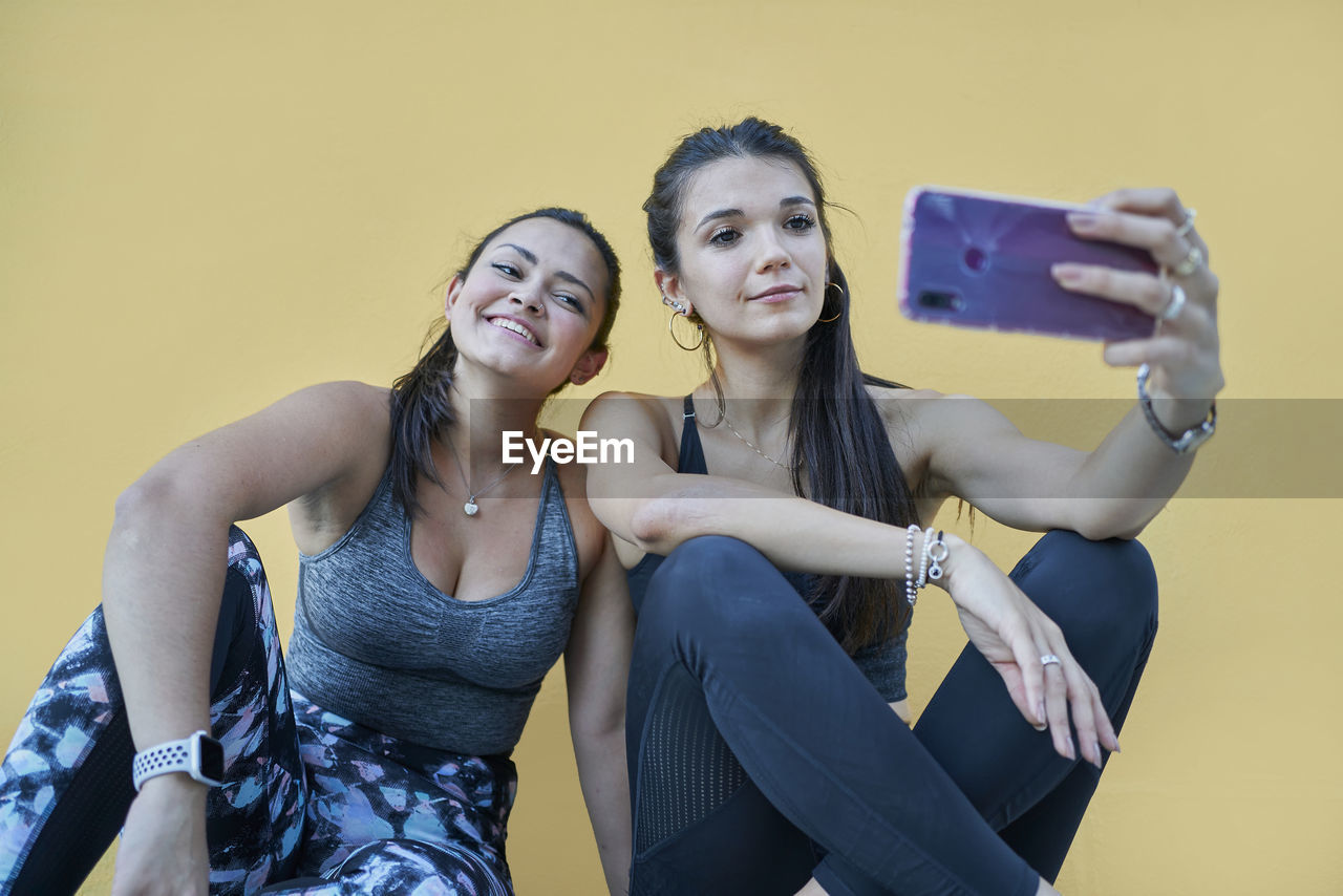 Full body content young female in activewear sitting on floor against dark yellow wall and taking selfies on modern mobile phone