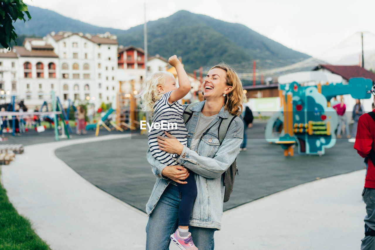 Happy laughing mother holding her little daughter in her arms is walking in the city.