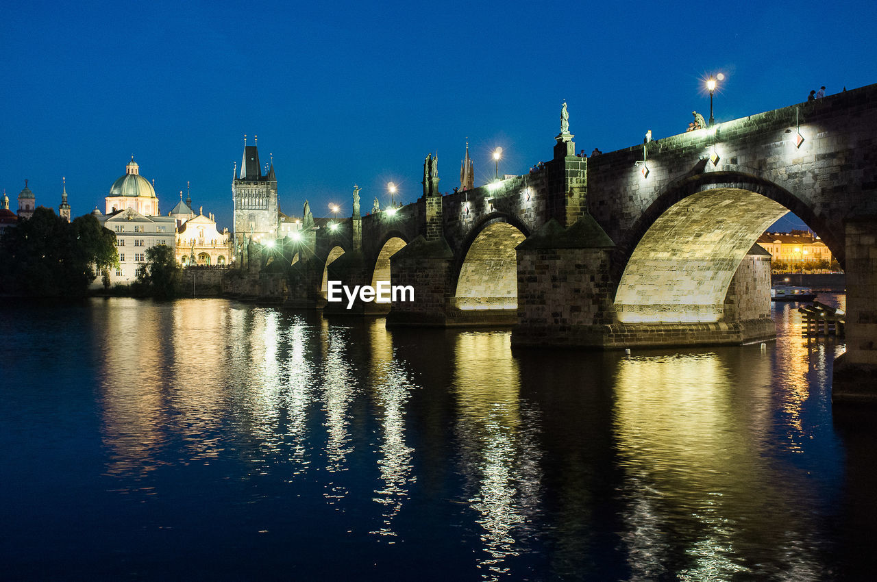 Illuminated arch bridge over river against blue sky at night