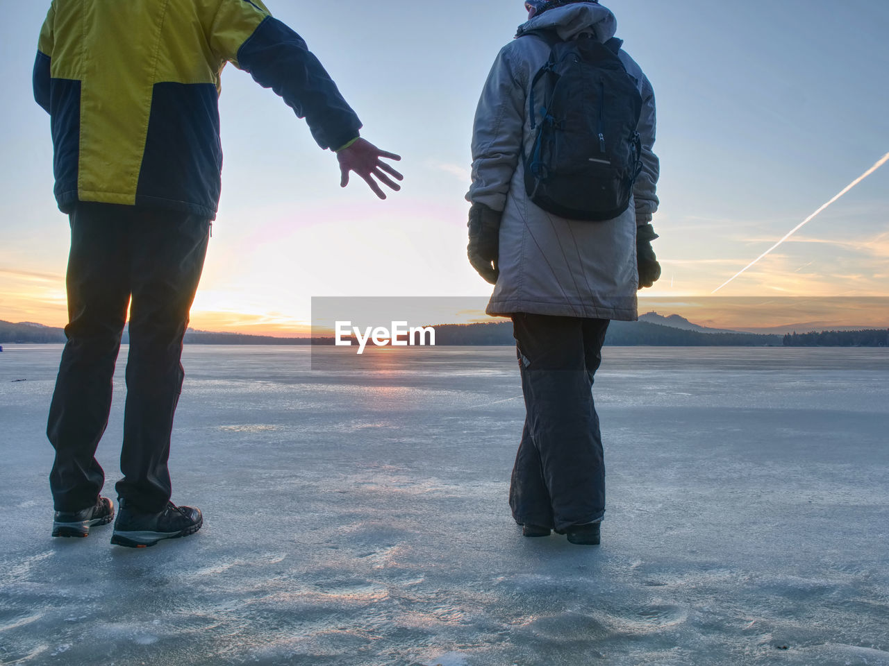 Tourist couple is spending time outdoors on a frozen lake, watching rising sun at horizon.