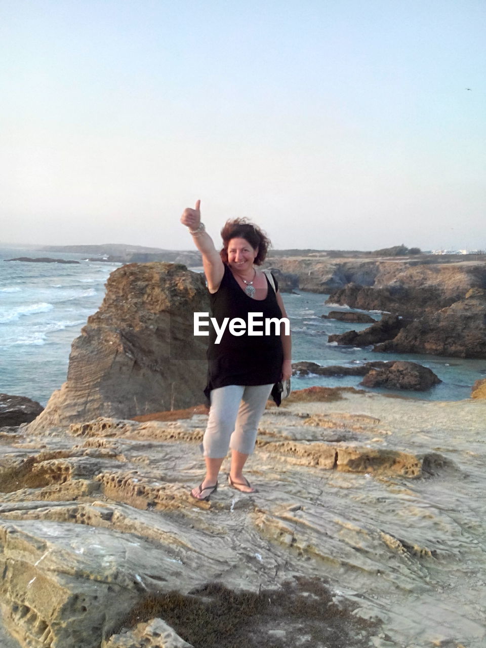 Portrait of woman showing thumbs up while standing on rock formation by sea against clear sky