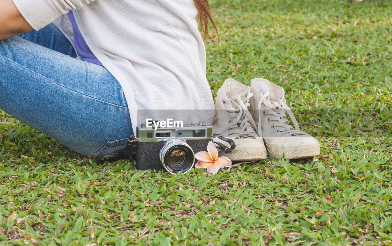 Midsection of woman sitting by vintage camera and sneakers on grassy field