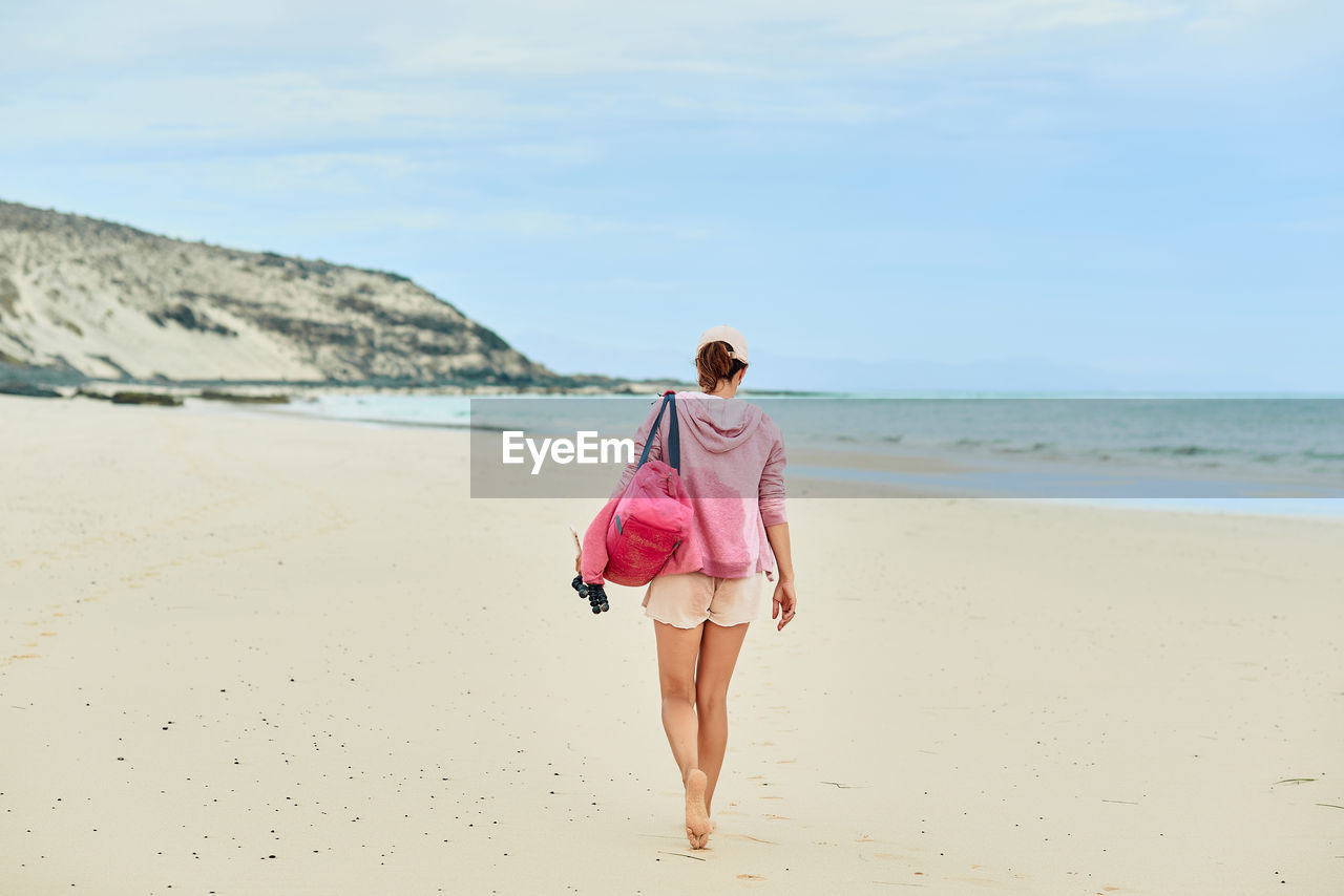 Back view full body of anonymous female tourist with bag walking barefoot along empty seashore towards waving sea