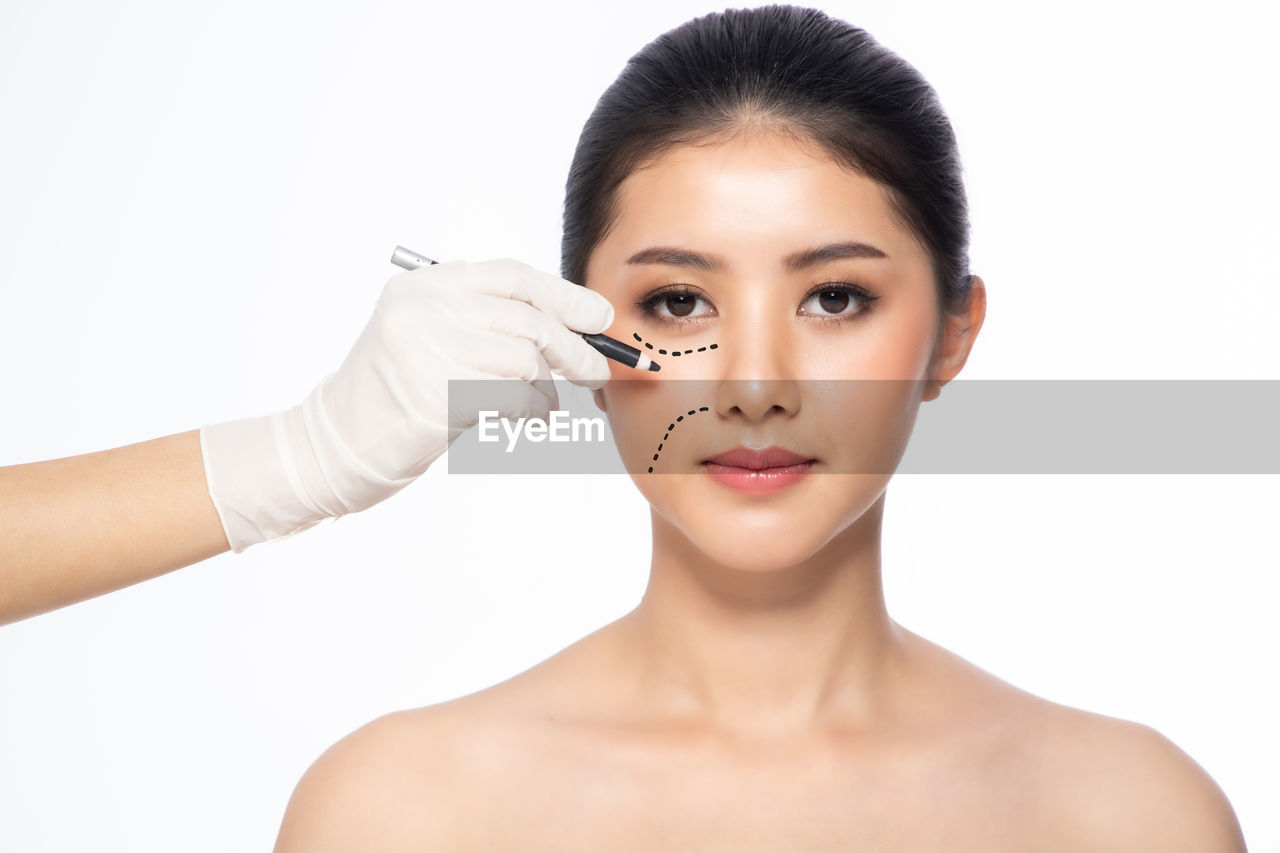 Cropped hands of doctor making markings on woman face against white background