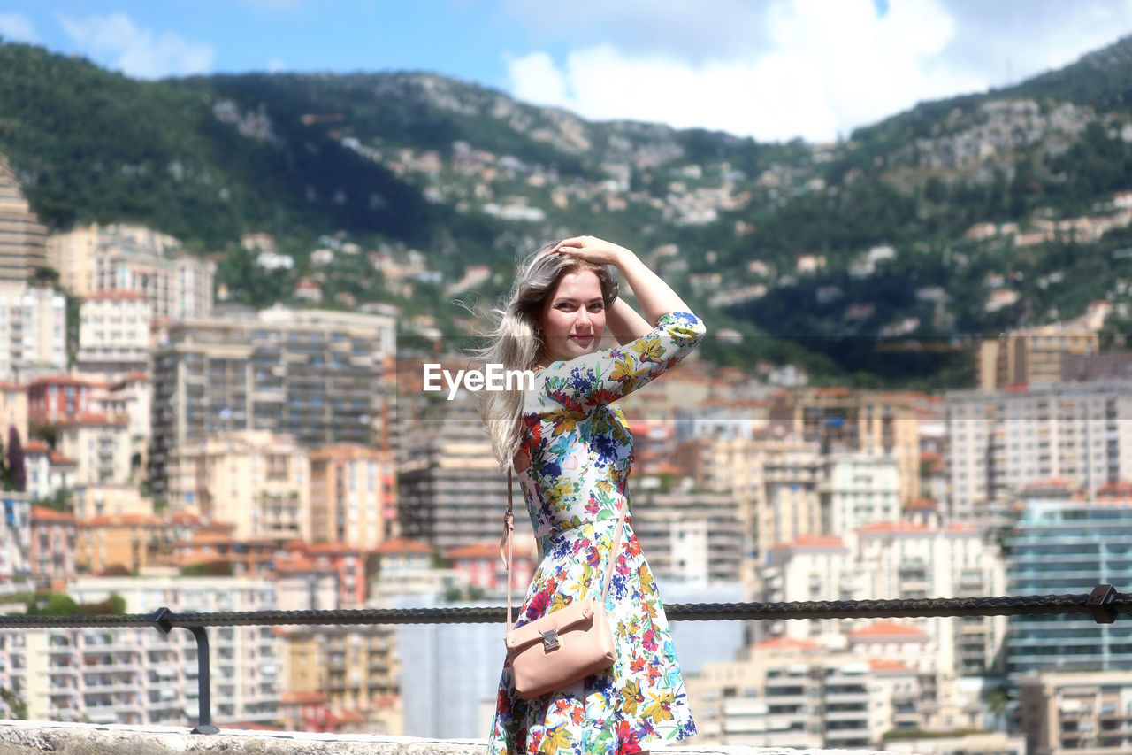 PORTRAIT OF YOUNG WOMAN STANDING AGAINST CITYSCAPE