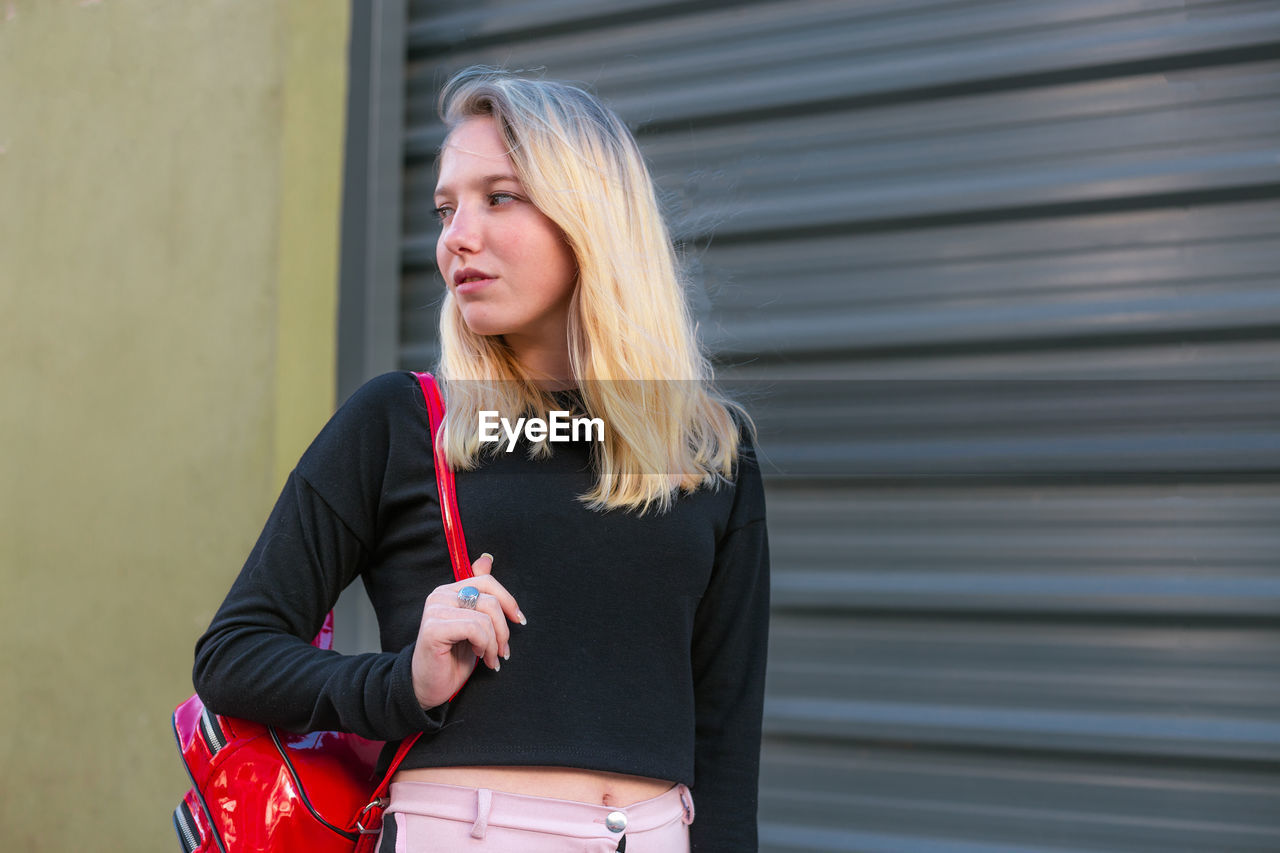 one person, women, adult, blond hair, red, hairstyle, lifestyles, clothing, standing, architecture, long hair, day, casual clothing, front view, female, looking, leisure activity, waist up, holding, young adult, person, outdoors, three quarter length, fashion, city, portrait, looking away, bag