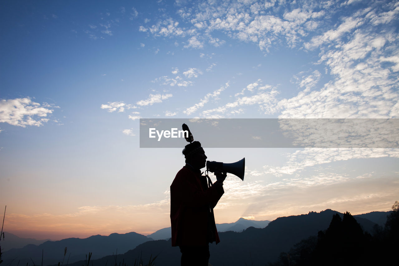 Silhouette man speaking on megaphone while standing against sky during sunset