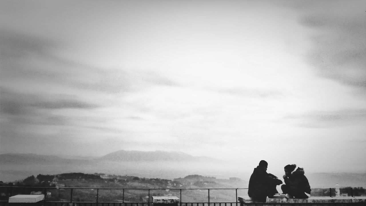 Man and woman sitting at observation point against cloudy sky