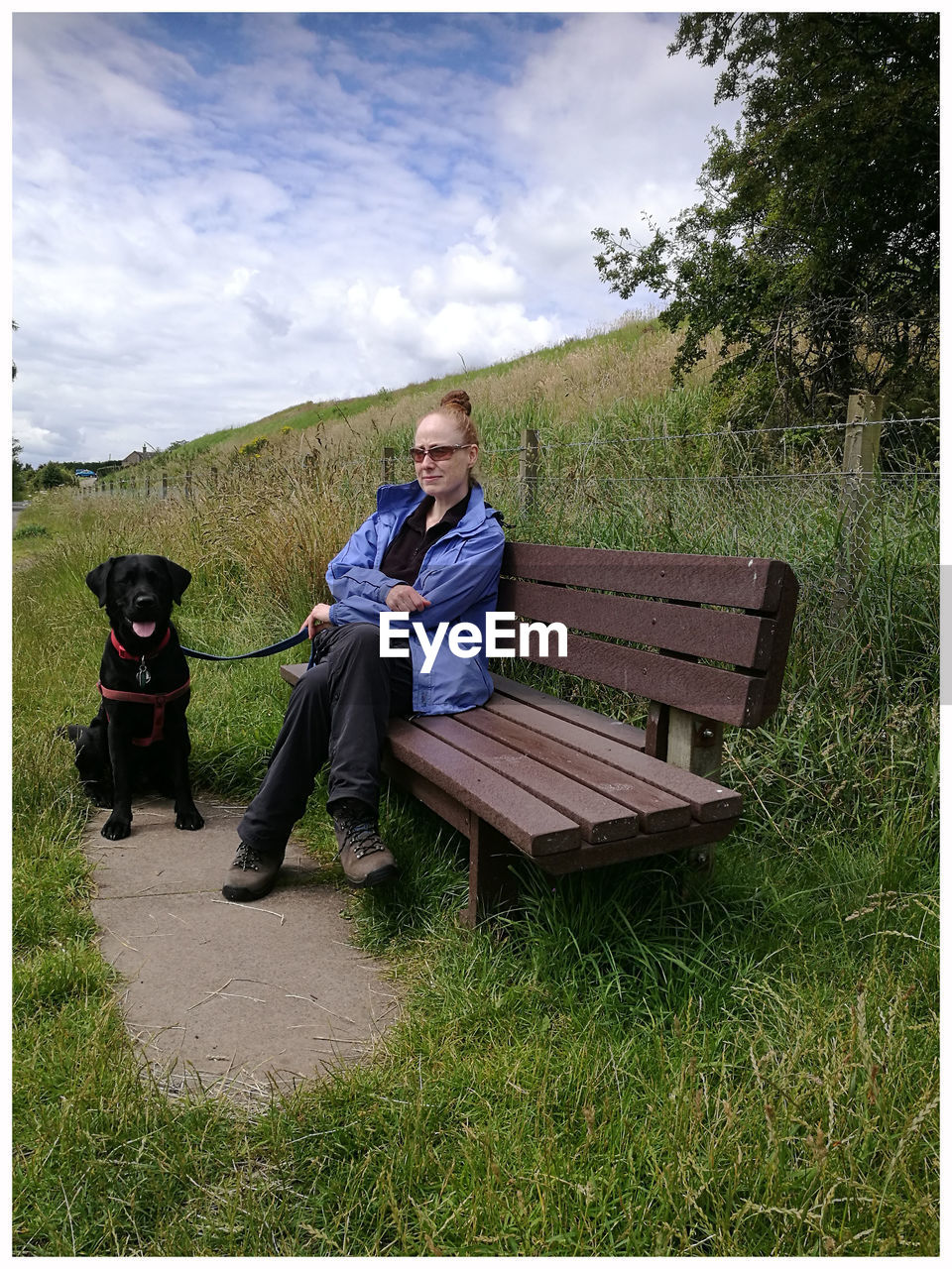 Mid adult woman with black labrador sitting on bench at park against cloudy sky