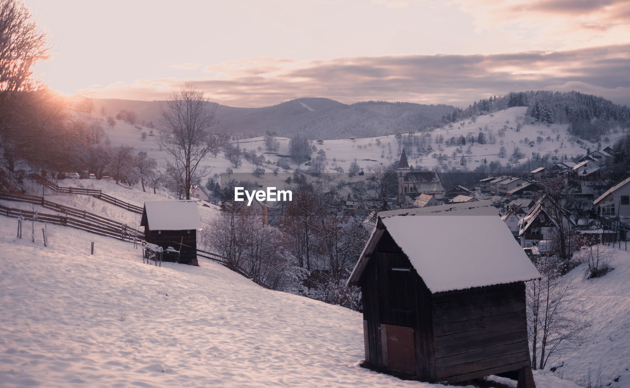 Wooden barns and cabind on snow covered field by buildings against mountains and sky