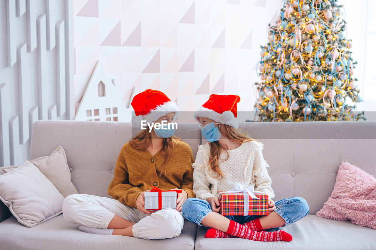 Full length sisters wearing mask sitting on sofa at home