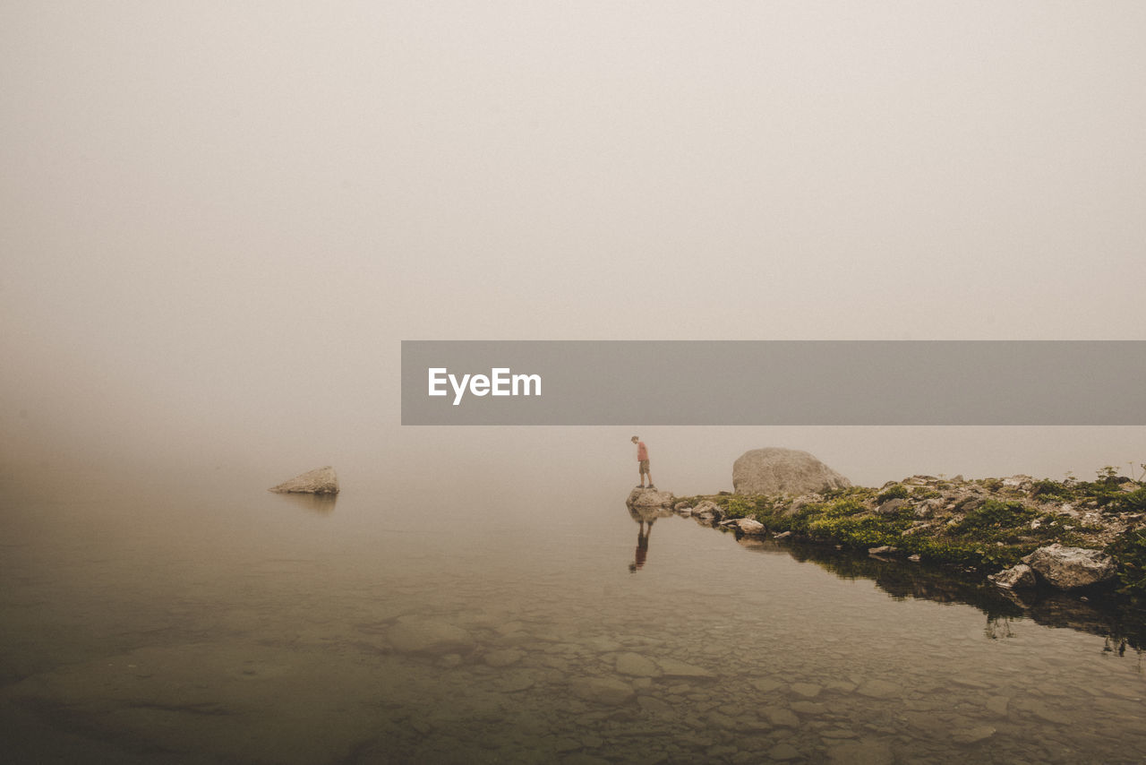 Distant view of man standing by sea during foggy weather