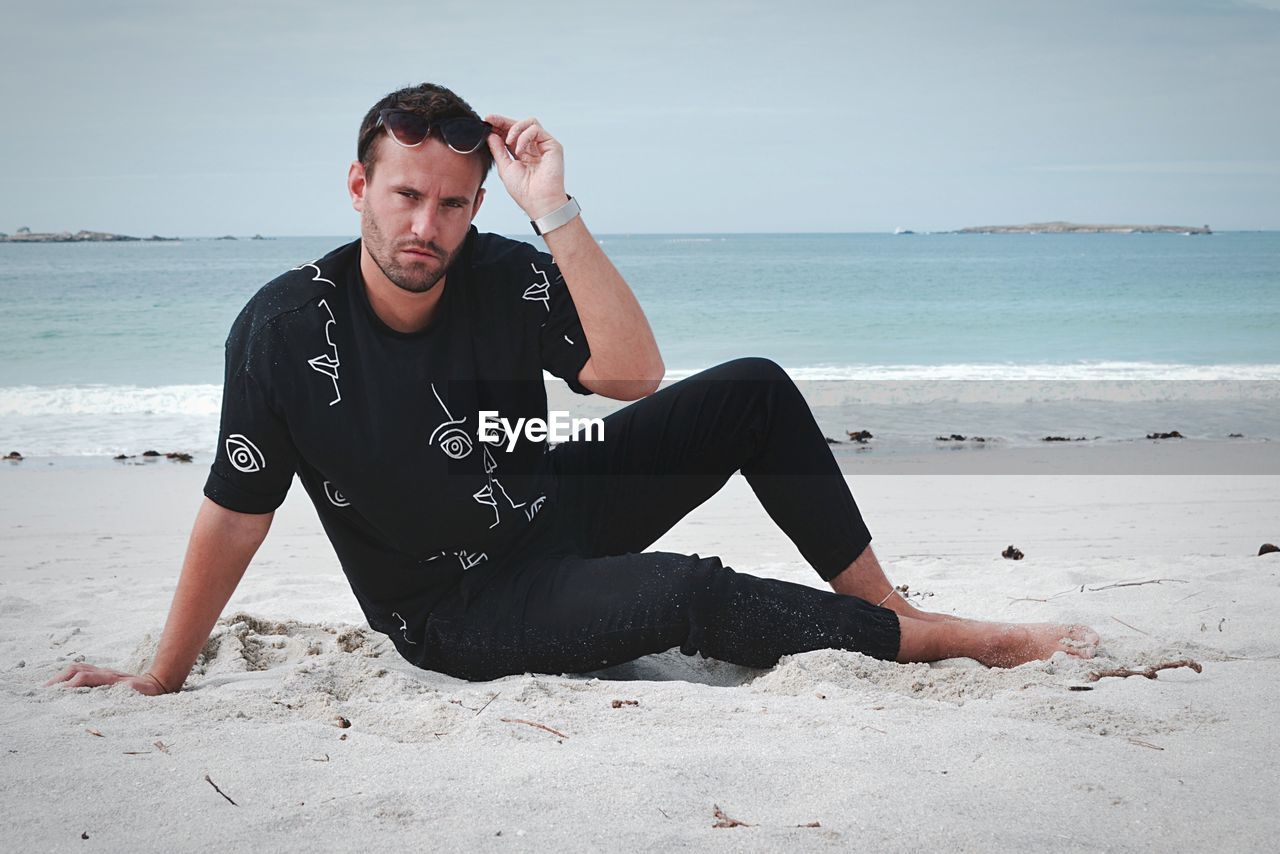 YOUNG MAN SITTING ON BEACH AGAINST SEA