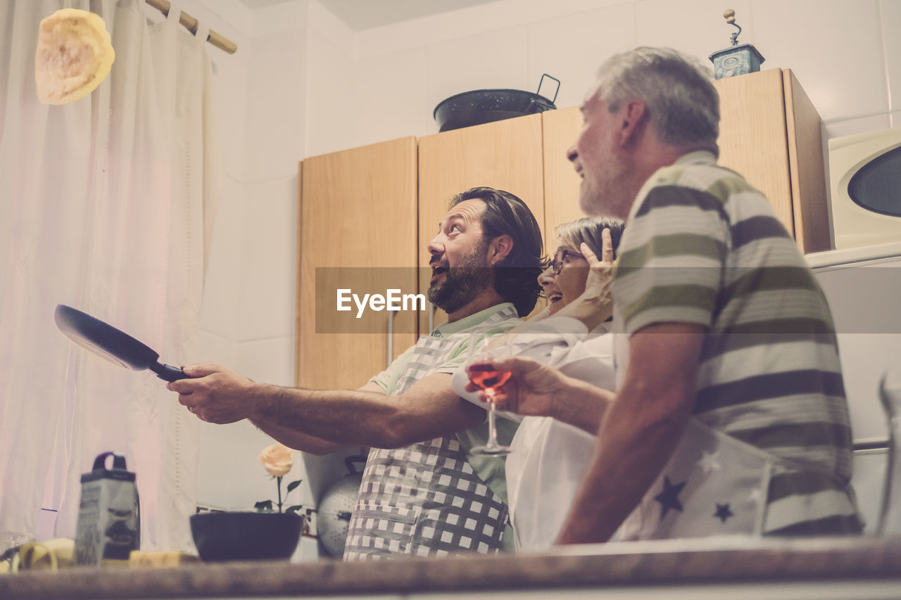 Mature son throwing omelette in pan while happy parents looking in kitchen