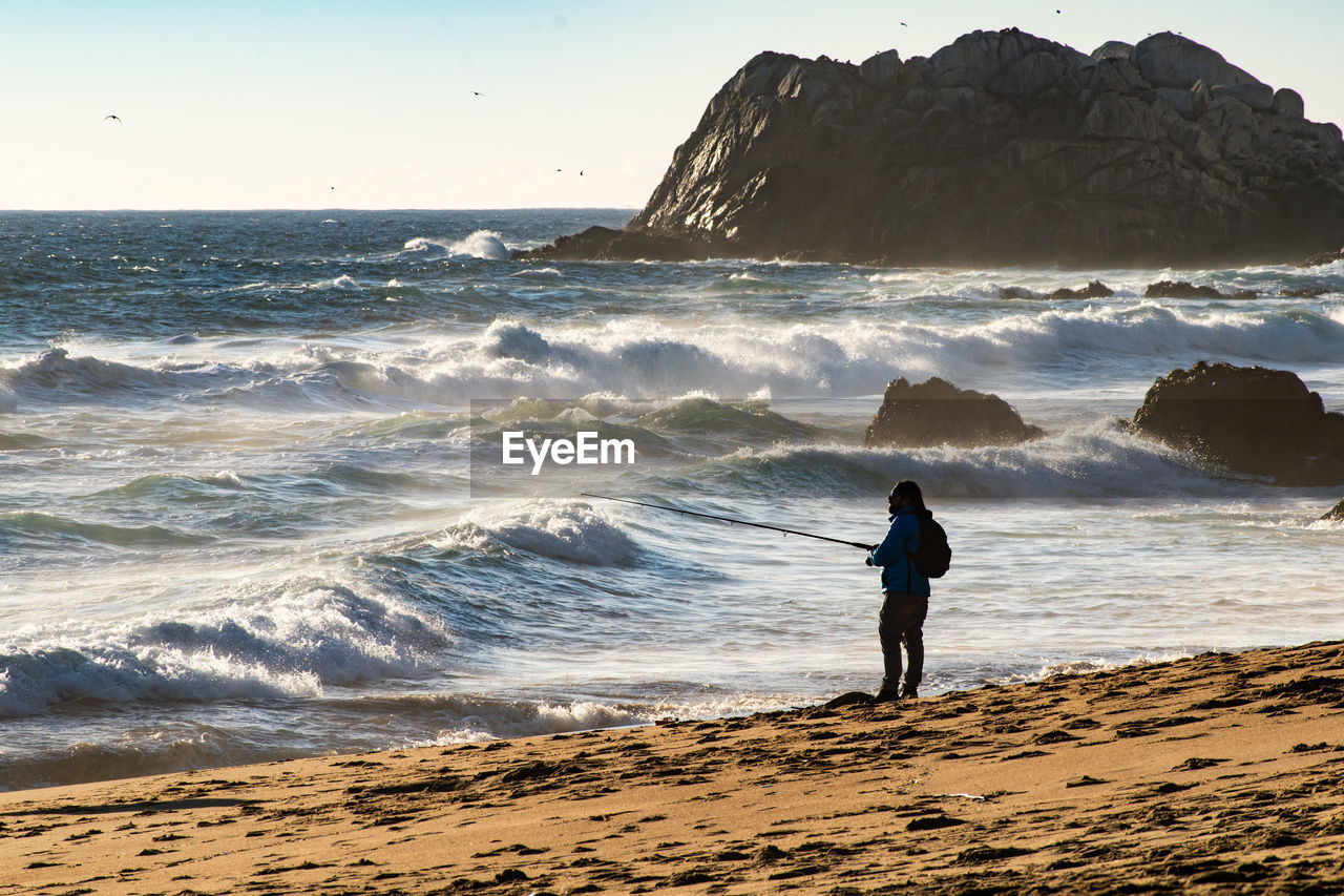 Full length of man fishing on beach against sky