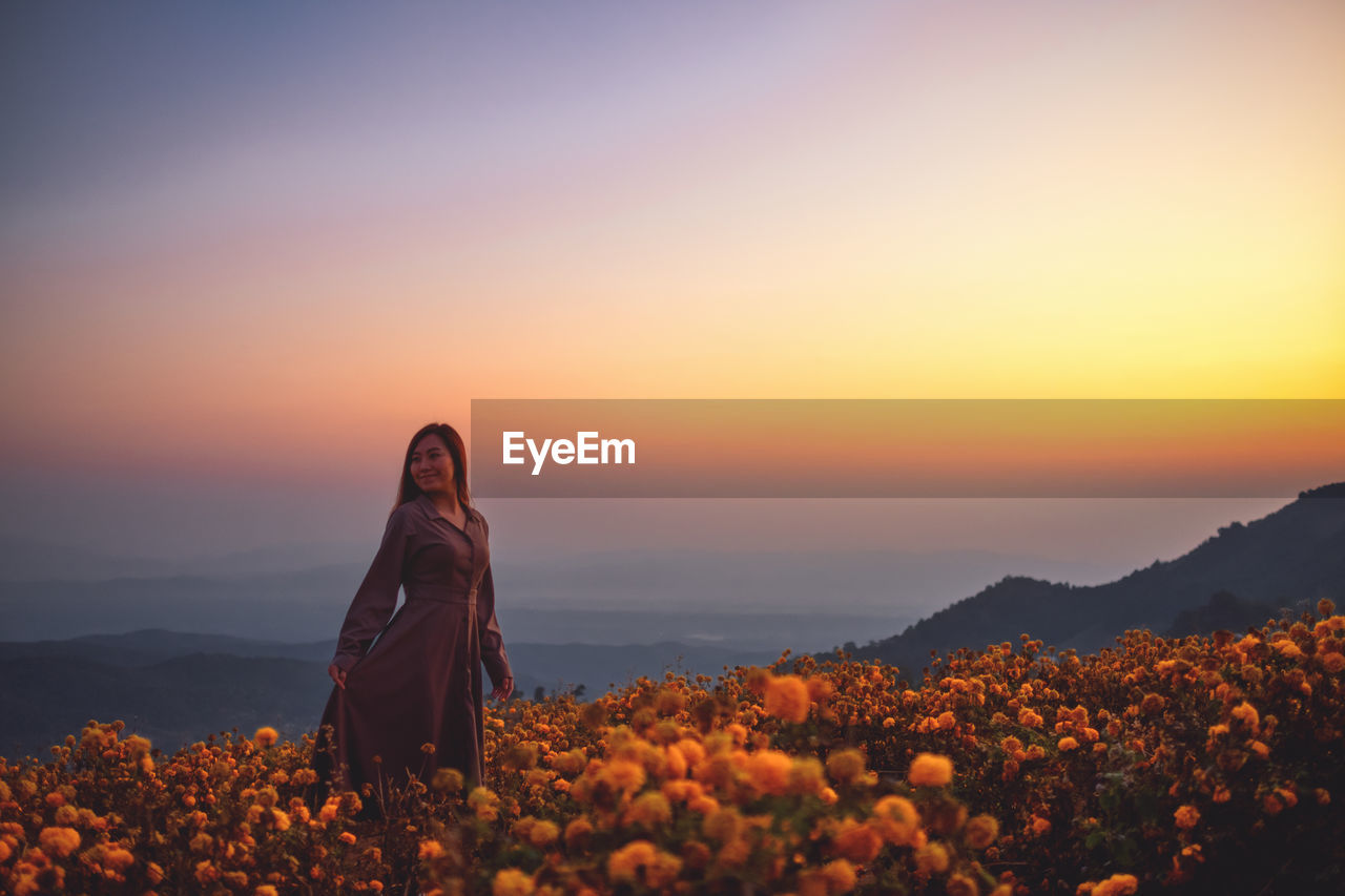 Woman standing by flowering plants against orange sky