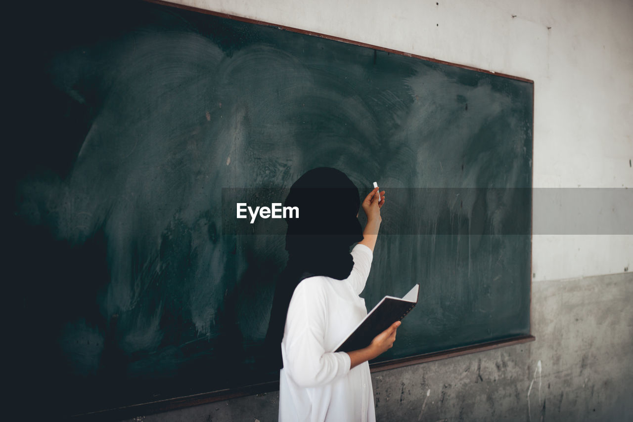 Teacher holding chalk while standing against blackboard in classroom