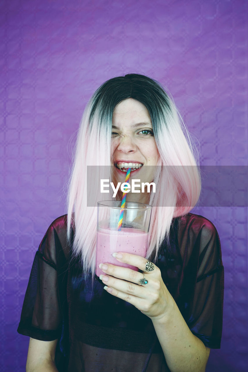 Portrait of young woman drinking smoothie while standing against wall