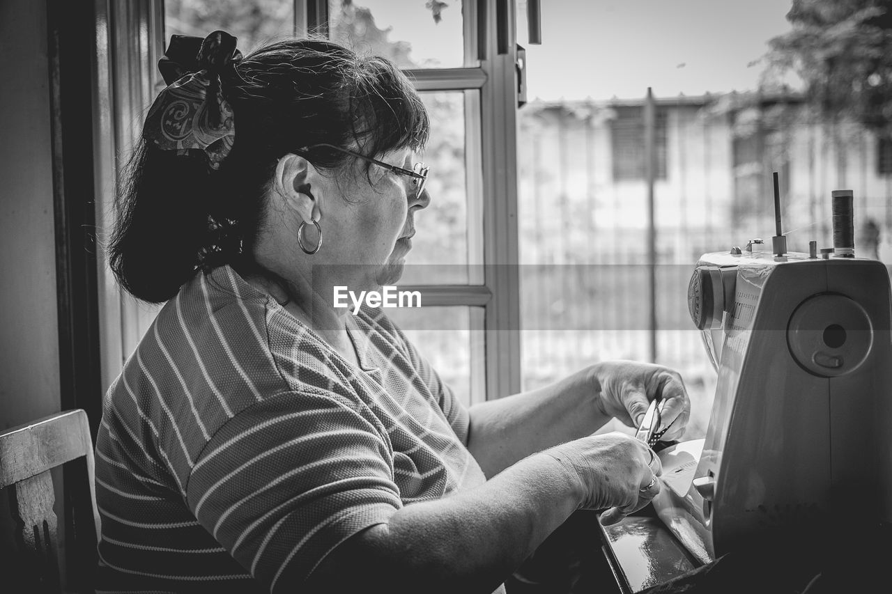 Side view of woman cutting textile while sitting with sewing machine at home