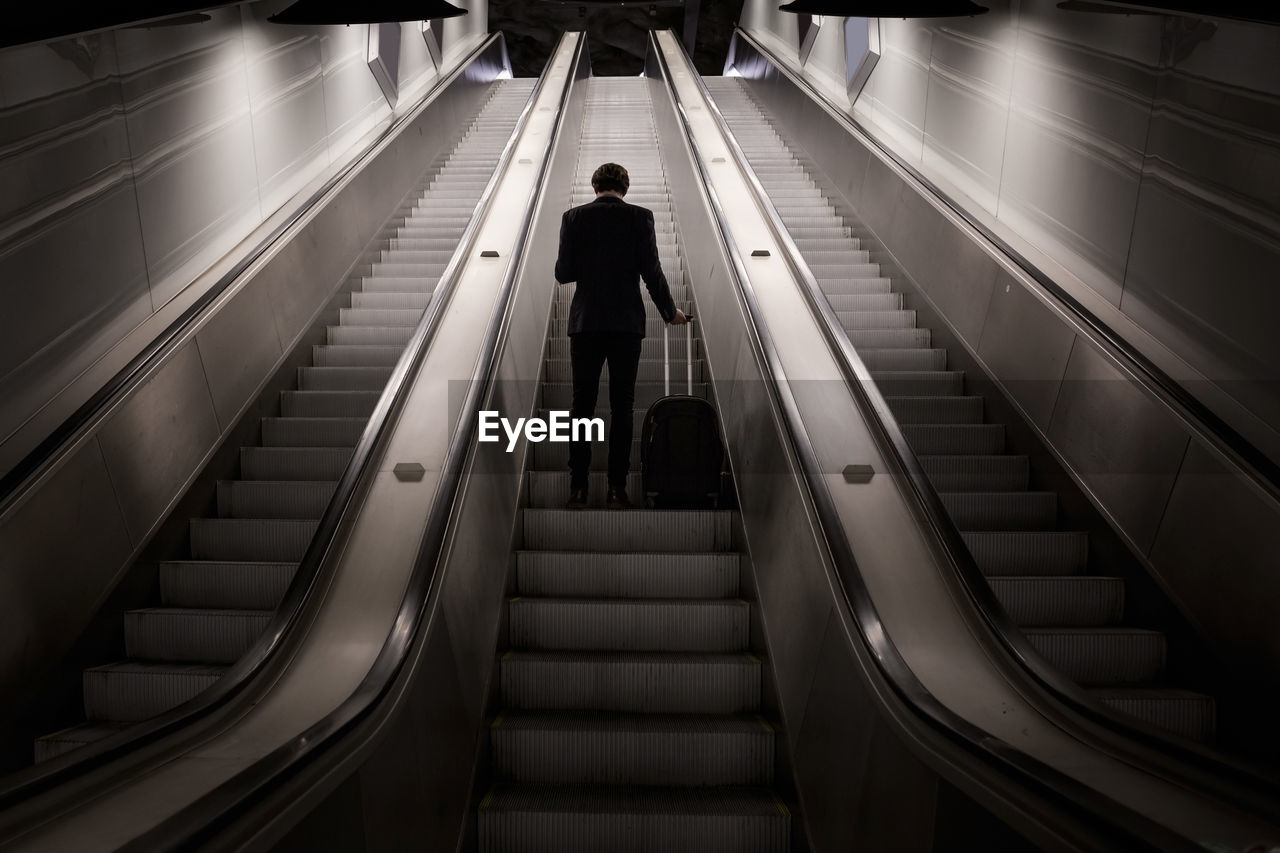 Low angle view of businessman standing with luggage on escalator in airport