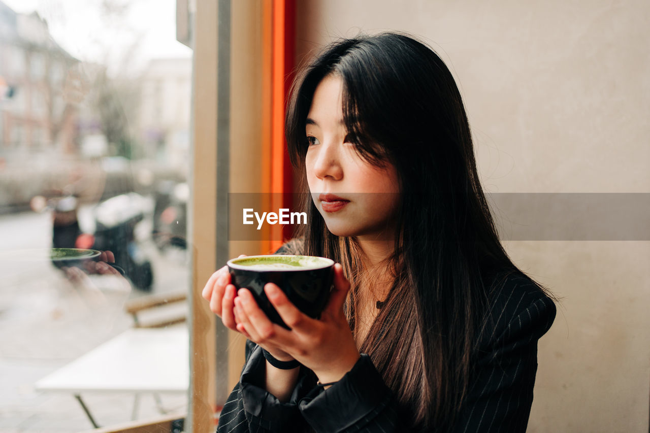 Long-haired brunette asian woman having a coffee on a coffee shop while is looking a cellphone through the window