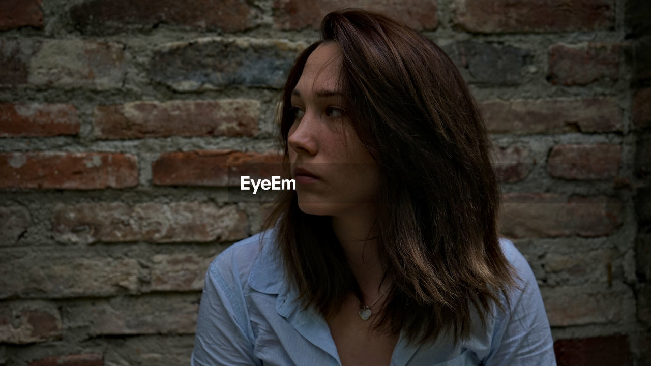 Close-up of thoughtful young woman looking away while standing against brick wall