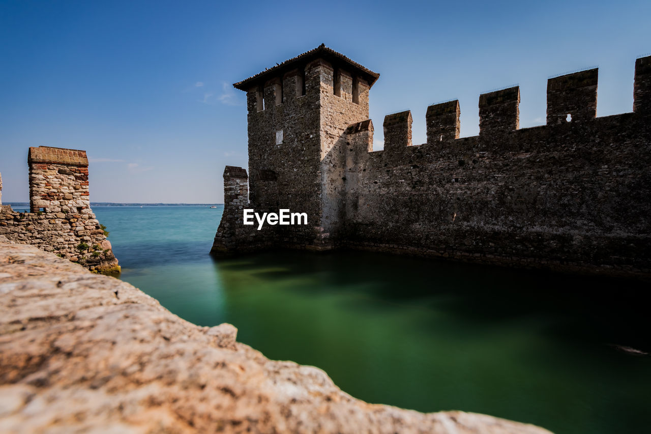 architecture, water, sky, built structure, history, sea, the past, castle, building exterior, nature, fort, rock, wall, travel destinations, coast, building, land, blue, reflection, fortification, clear sky, travel, fortified wall, beach, medieval, tower, old ruin, no people, ancient, old, ocean, stone material, outdoors, scenics - nature, moat, tourism, landmark, terrain, coastline, ancient history, cliff, ruins, surrounding wall