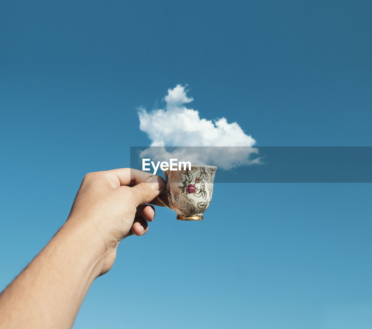 Optical illusion of cropped hand holding cup with cloud against blue sky