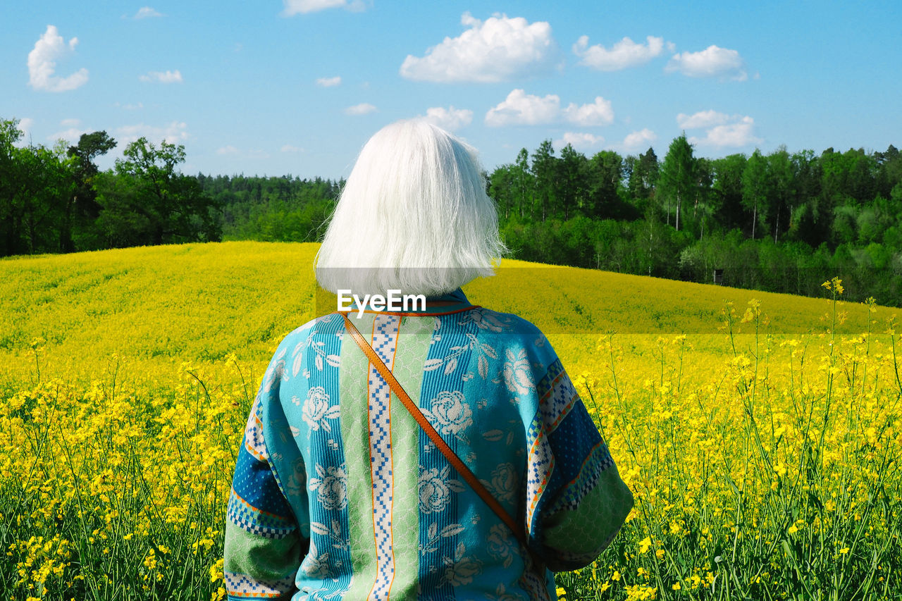 Rear view of senior woman standing on rapeseed field against sky