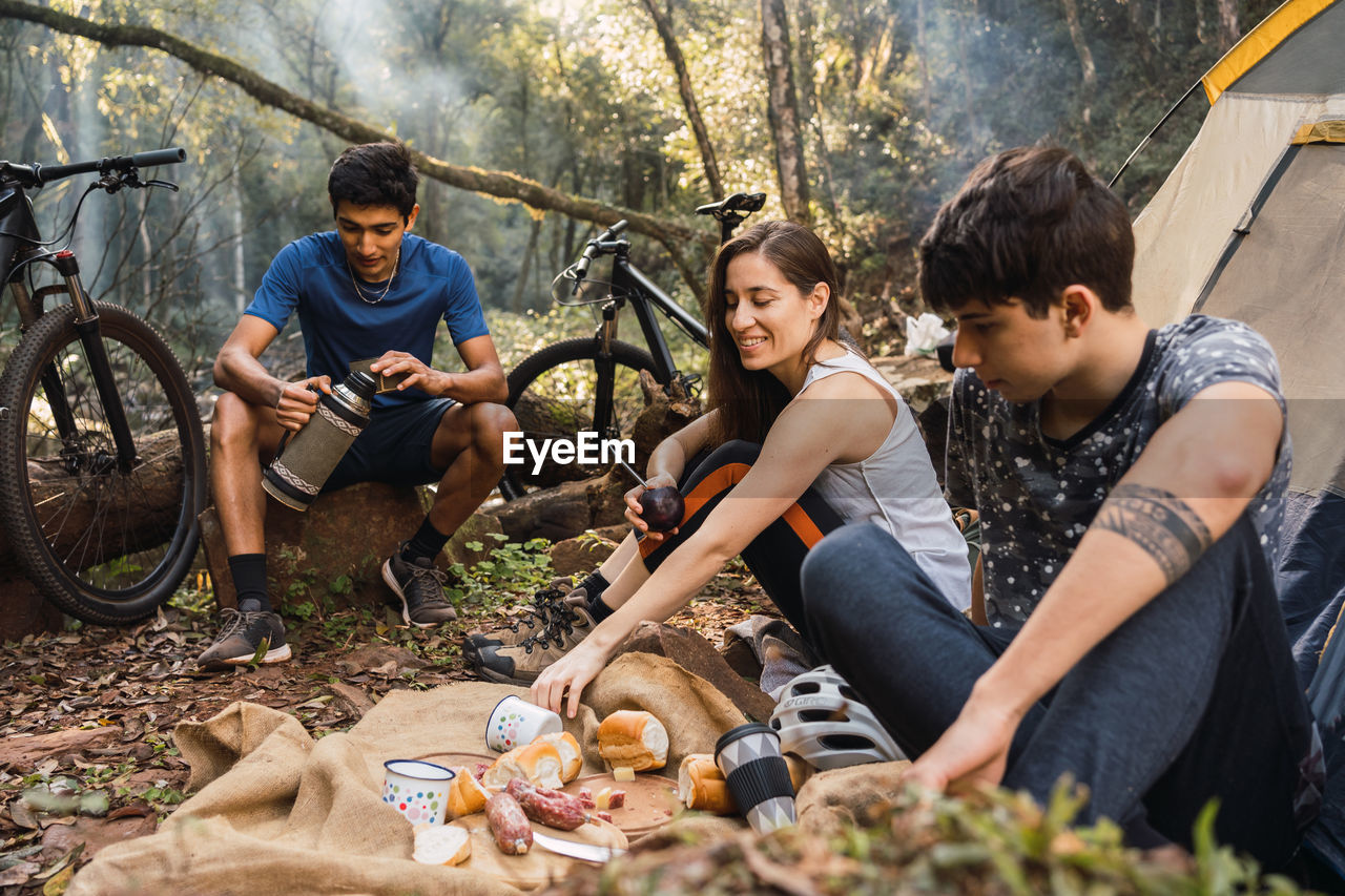 Diverse cyclists sitting on ground near tent and having picnic while hiking together in forest