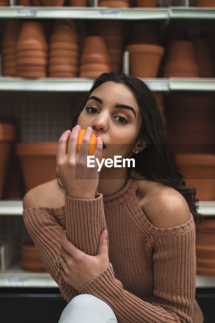 Portrait of teenage girl eating orange against flower pots on shelves in store