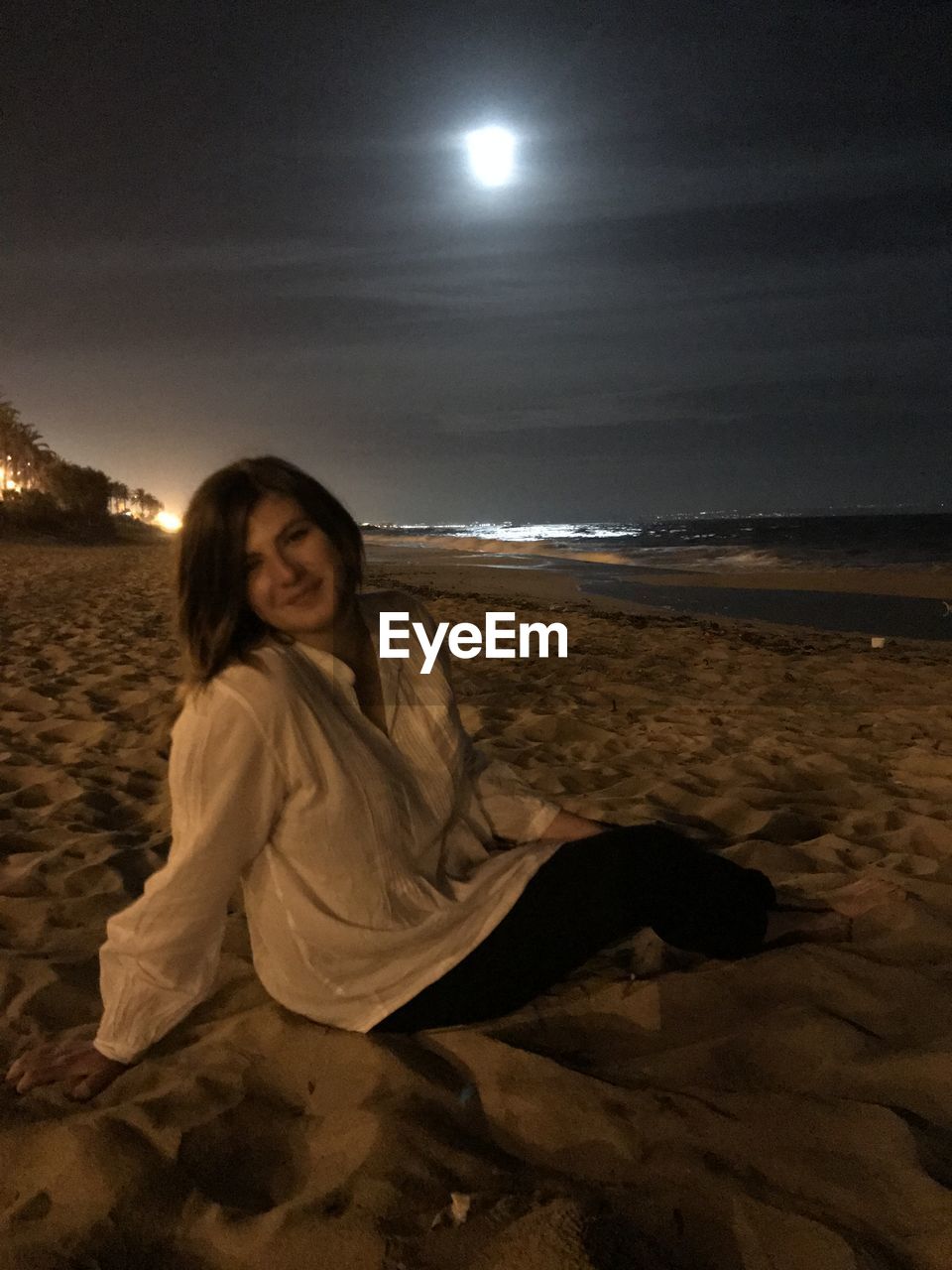 BEAUTIFUL YOUNG WOMAN SITTING ON BEACH AGAINST SKY AT NIGHT