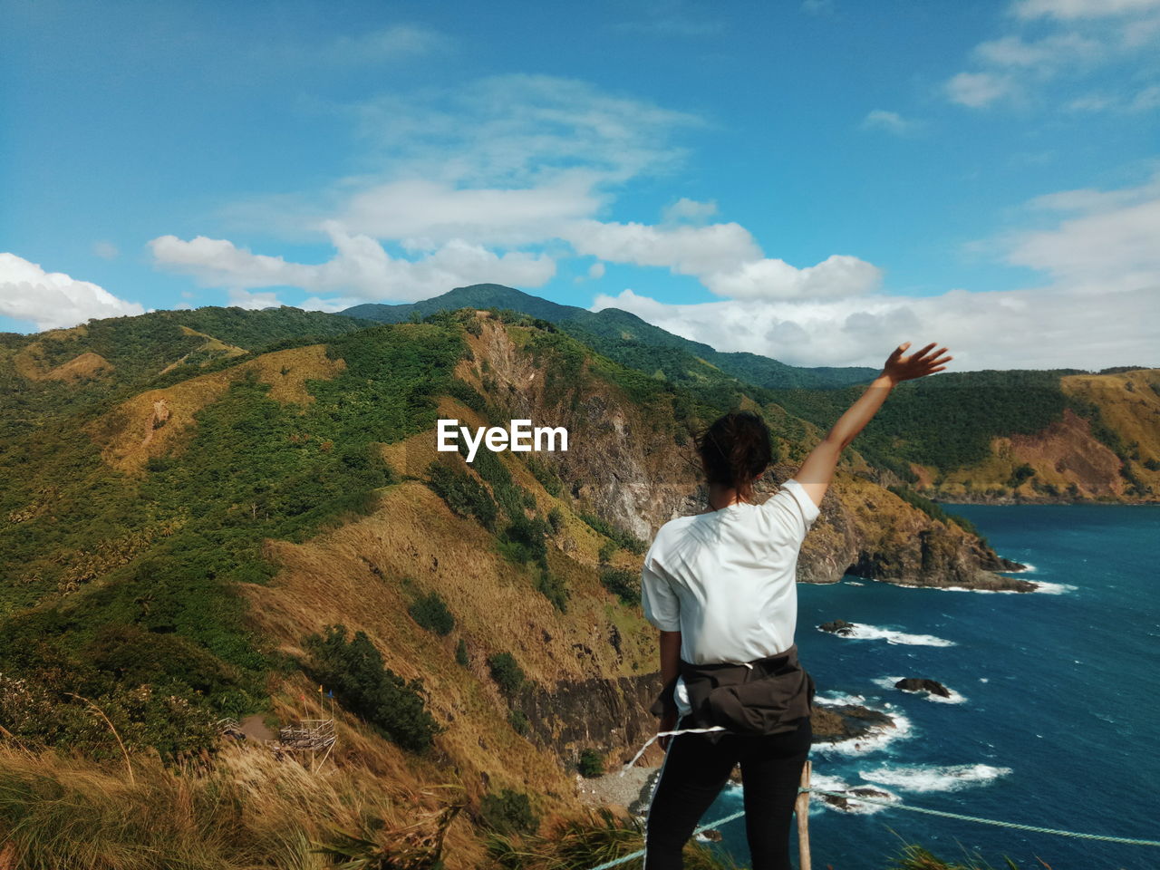 Rear view of woman waving hand while standing on cliff by sea against sky