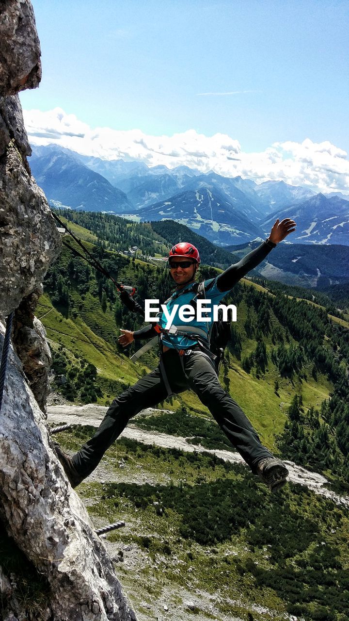 Carefree man hanging from harness on cliff against mountains