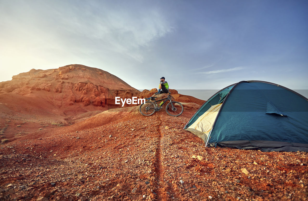 Mid distance view of hiker sitting by mountain bike on rocks at desert with tent in foreground